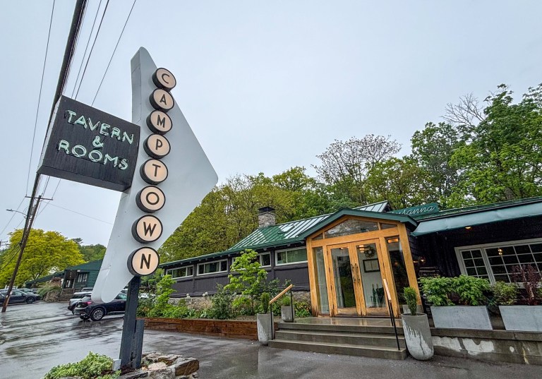 Retro roadside sign with vertical letters spelling CAMP TOWN and a dark panel reading TAVERN & ROOMS beside a green-roofed storefront and trees behind it.